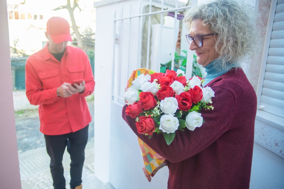 A woman with curly gray hair, wearing glasses, a blue turtleneck, and a maroon cardigan, holds a vibrant floral gift bouquet featuring red and white roses, wrapped in colorful patterned paper. She sta