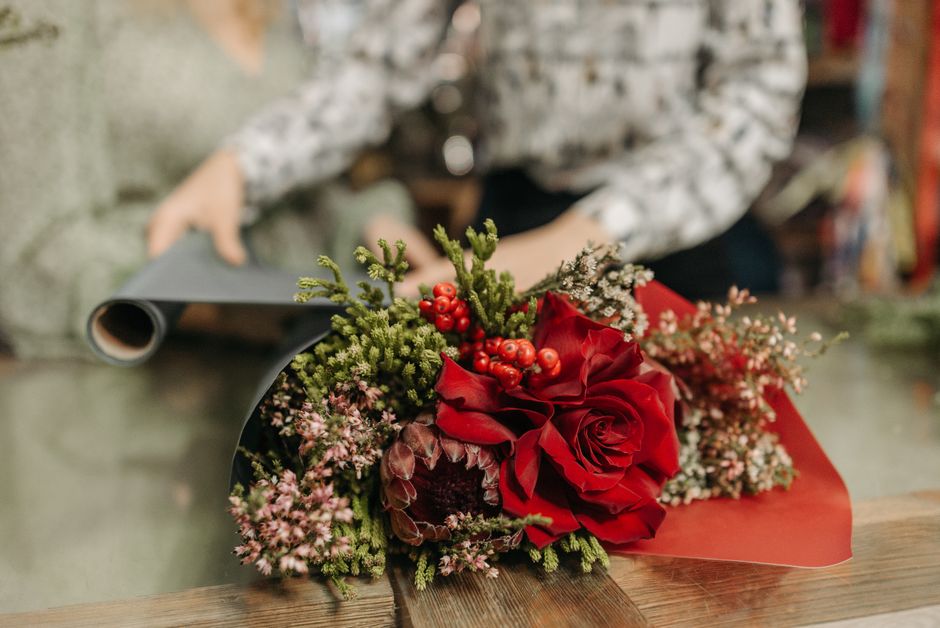 A floral bouquet featuring a large, deep red rose at the center, surrounded by sprigs of small pink and white flowers, green foliage including pine-like needles, and clusters of bright red berries. Th