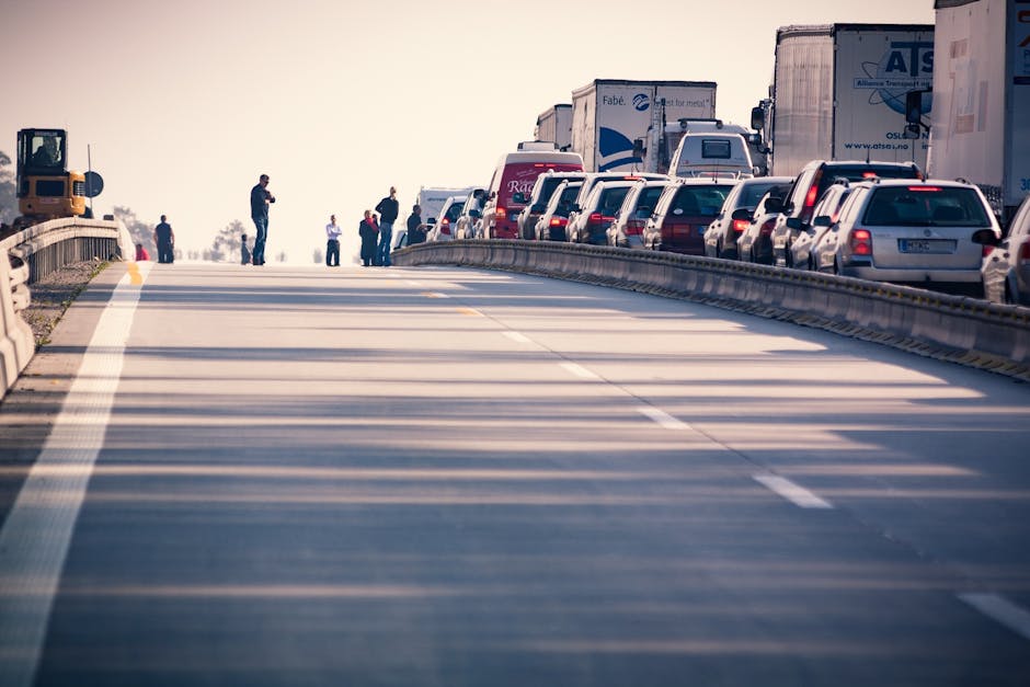 A busy highway with multiple lanes filled with lined-up vehicles including cars and trucks, extending towards the horizon. Several pedestrians are seen standing on the side of the road near the cars,