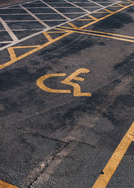 A close-up view of a disabled parking space marked with yellow lines on dark asphalt, featuring a yellow wheelchair accessibility symbol painted directly onto the surface. The parking area appears emp