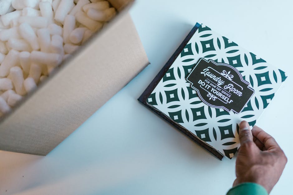A person's hand holding the corner of a black and white patterned box with a label that reads 'Laundry Goom for Same Day Service Do It Yourself', positioned on a white surface. To the left, part of a
